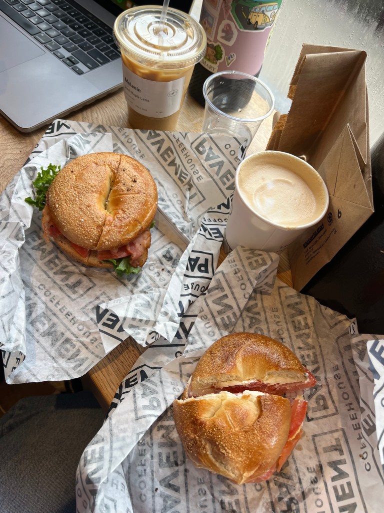 Two bagel sandwiches, a paper cup with a hot latte, and a plastic cup with iced coffee in it on a crowded table.
