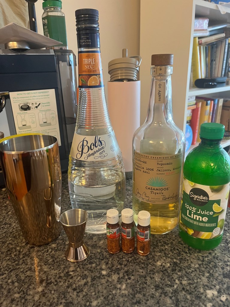 A cocktail shaker, a couple bottles of liquor, a bottle of lime juice, and mini bottles of Tajin set out on a counter.