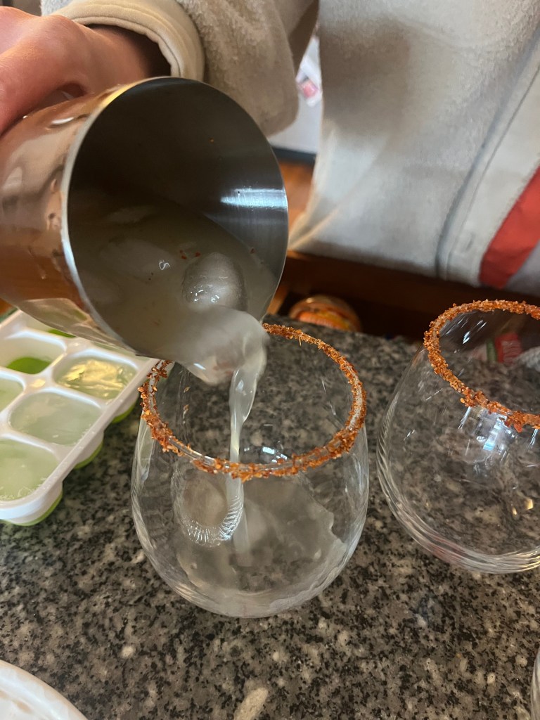 A drink being poured from a cocktail shaker into a rimmed glass.